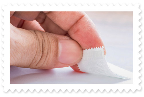 A colorful photograph featuring a closeup of a hand placing an adhesive stamp in the corner of an envelope.